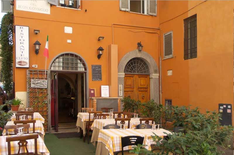 Dehors con tavoli apparecchiati a quadretti gialli e bianchi davanti a una facciata color arancio con due portali ad arco.