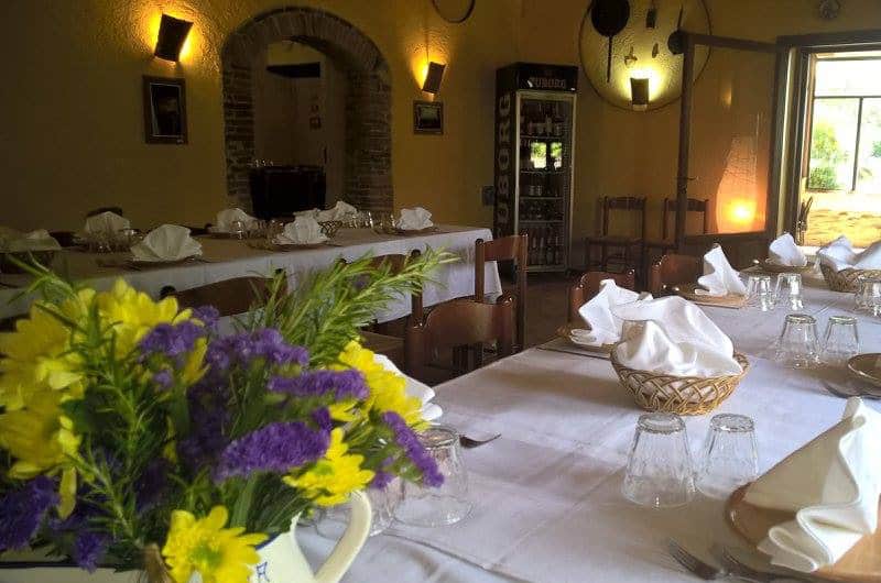 Tavoli con tovaglie bianche e cestini di tovaglioli, in primo piano un vaso di fiori gialli e viola.