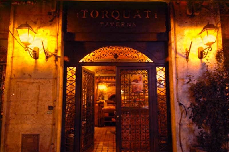 Ingresso serale con insegna Torquati Taverna, due lanterne accese e porta in ferro battuto aperta sulla sala illuminata.