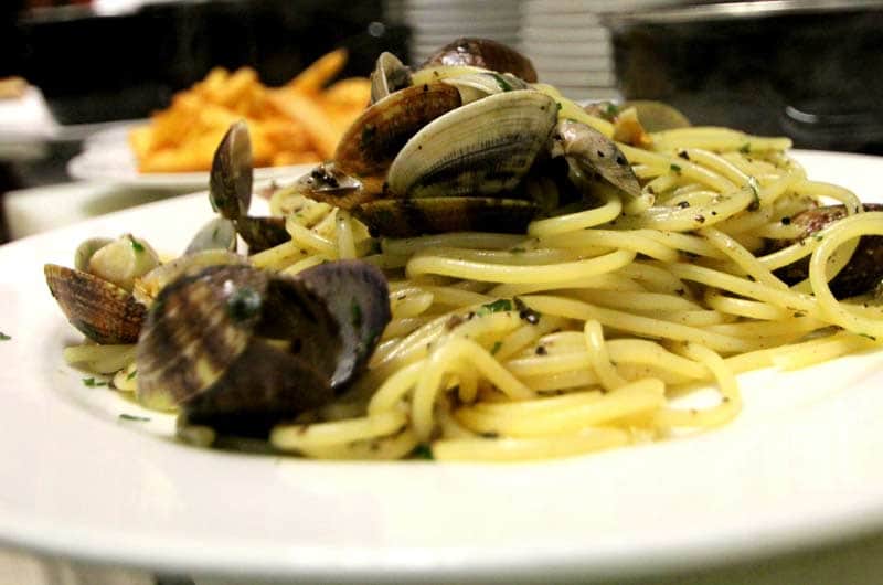 Spaghetti con vongole in guscio aperte, cosparsi di prezzemolo tritato e pepe nero, su piatto bianco.