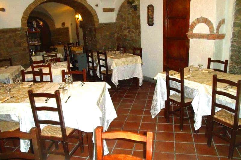 Sala con volte in pietra e mattoni, tavoli quadrati con tovaglie bianche e sedie in legno scuro.