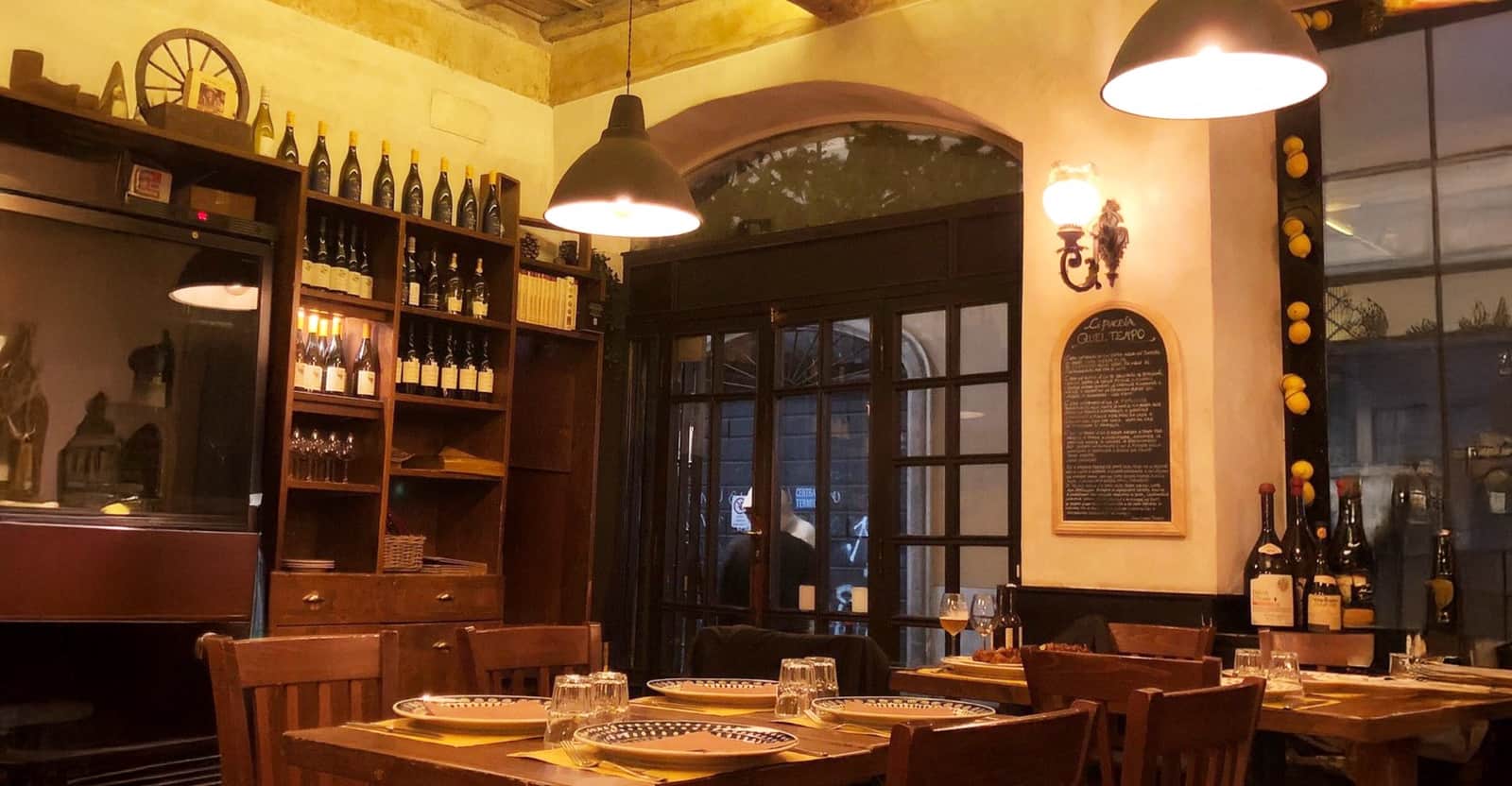 Sala con tavoli in legno apparecchiati, scaffali pieni di bottiglie di vino e due lampade a cupola nera accese.