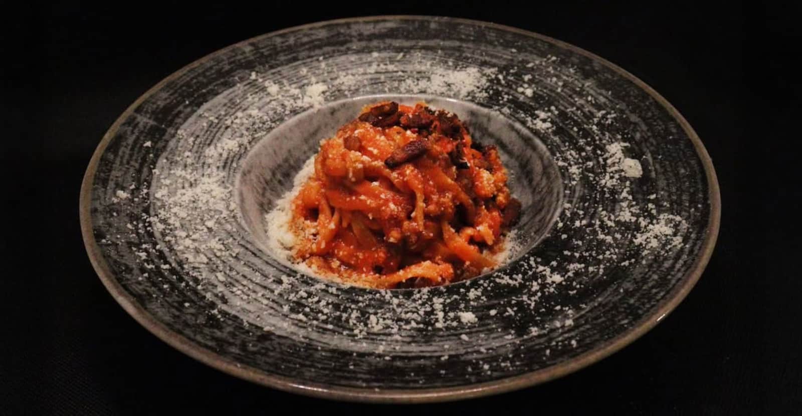 Spaghetti al sugo di pomodoro con pezzetti scuri e formaggio grattugiato, in un piatto largo grigio scuro.