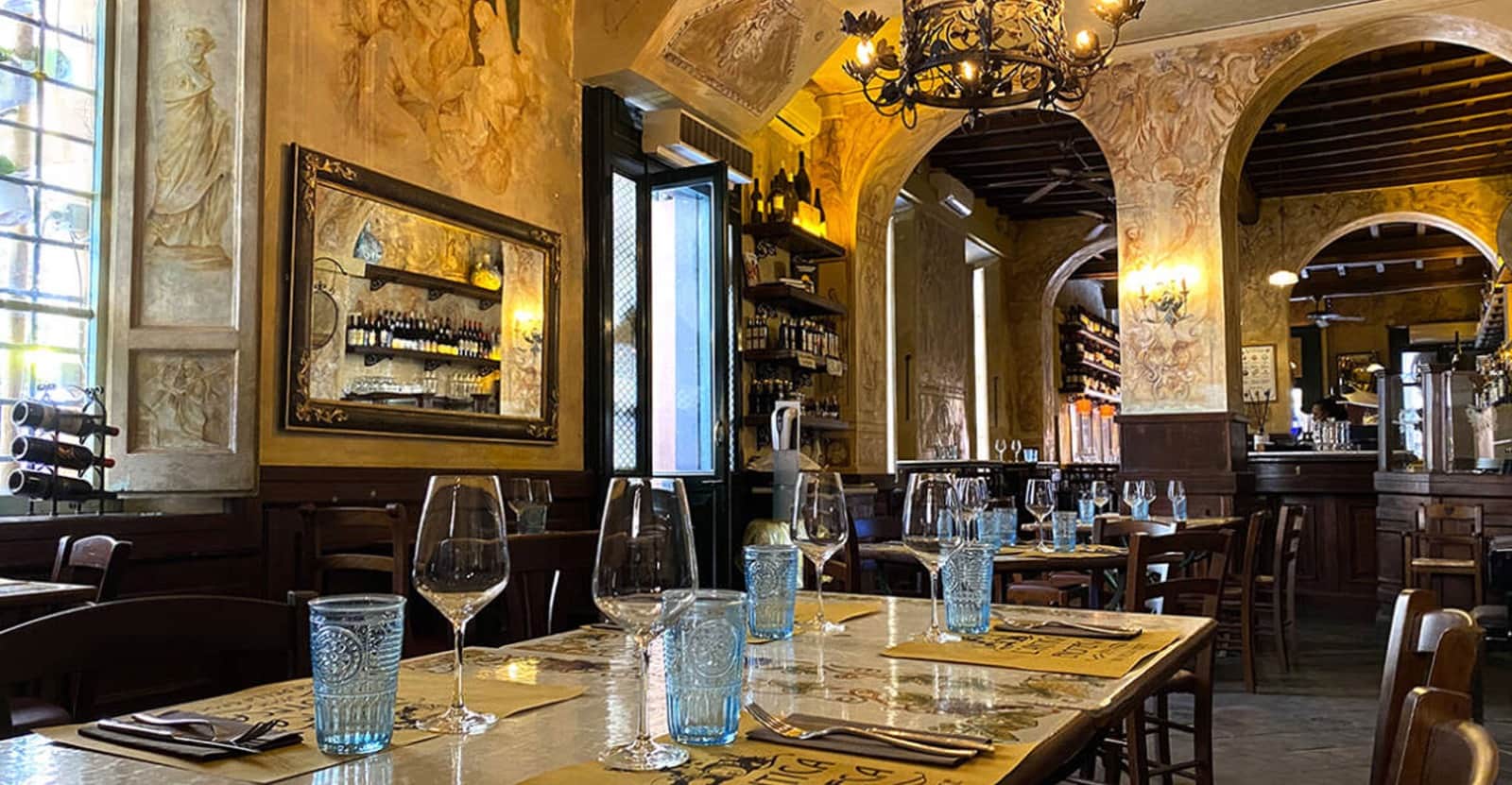 Sala con pareti affrescate, archi in pietra e tavoli apparecchiati con bicchieri azzurri e calici da vino.