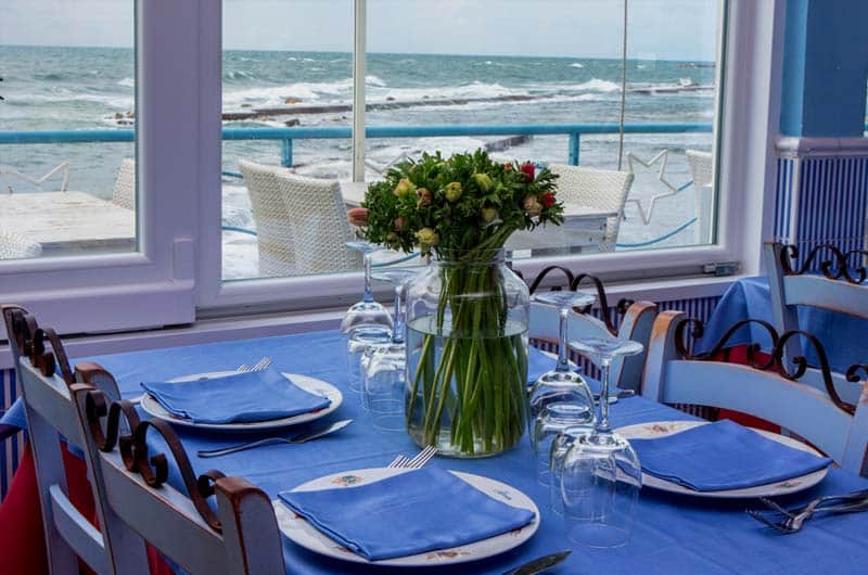 Tavolo con tovaglia blu, bicchieri e piatti apparecchiati davanti a vetrate affacciate sul mare mosso.