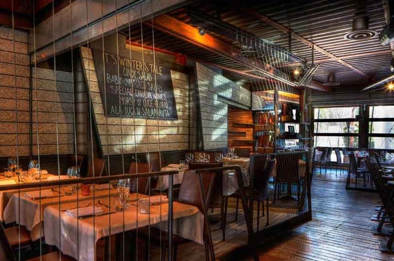 Sala di ristorante con tavoli apparecchiati, cavi metallici a vista e una lavagna con menù scritto a gesso.