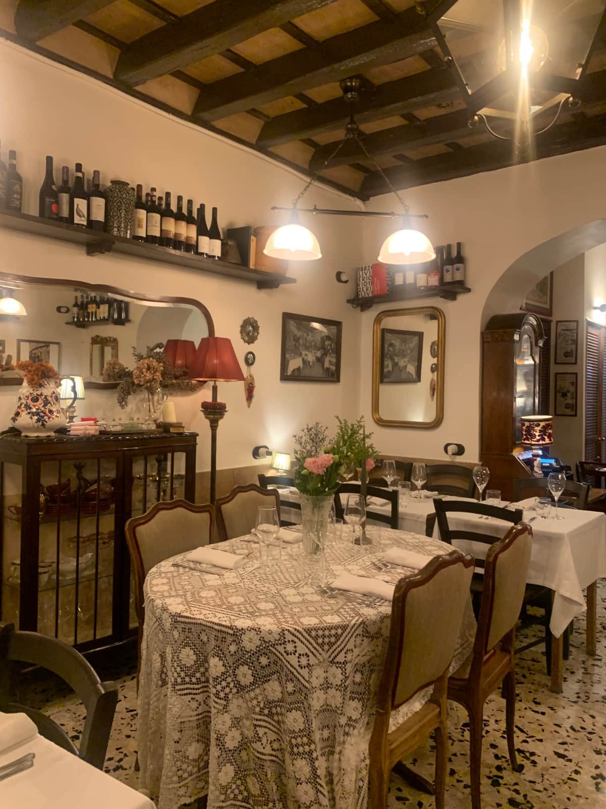 Sala con travi a vista, tavoli apparecchiati con tovaglie di pizzo e fiori rosa al centro.