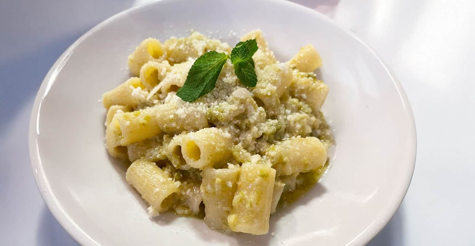 Rigatoni conditi con sugo verde e formaggio grattugiato, decorati con due foglie di menta, in un piatto bianco.
