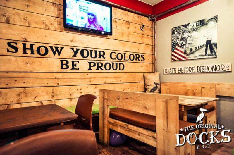 Parete in legno con scritta "Show your colors, be proud", televisore a muro e sedute imbottite marroni.
