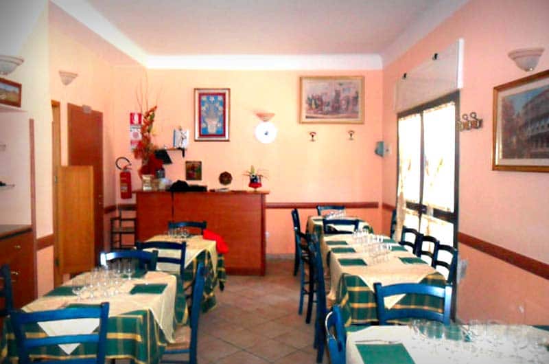 Sala da ristorante con tavoli quadrati apparecchiati con tovaglie a quadri verdi e sedie blu in legno.