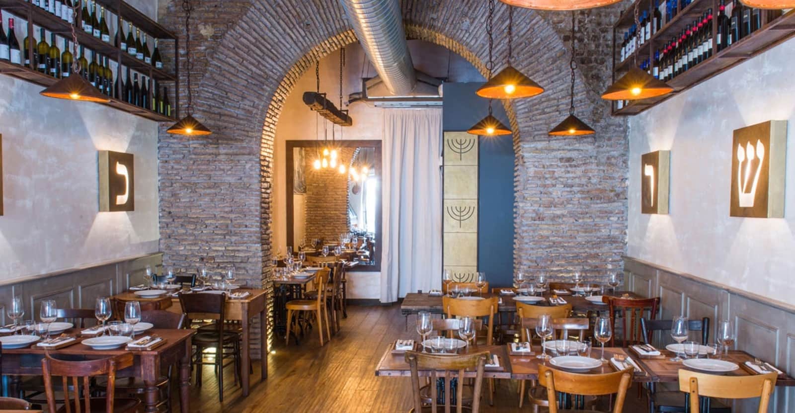 Sala da ristorante con soffitto a volta in mattoni, tavoli in legno apparecchiati e lampade a sospensione dai paralumi ramati.