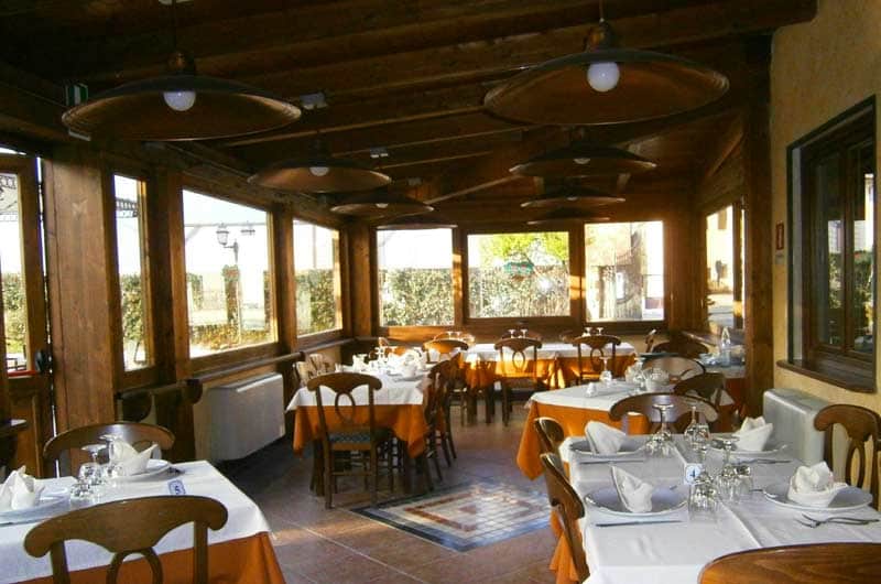 Sala da pranzo con travi in legno a vista, tavoli apparecchiati con tovaglie bianche e arancioni e grandi lampade a sospensione.
