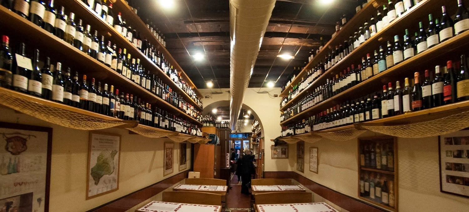 Corridoio con scaffali di legno colmi di bottiglie di vino su entrambi i lati, tavolini apparecchiati in primo piano.