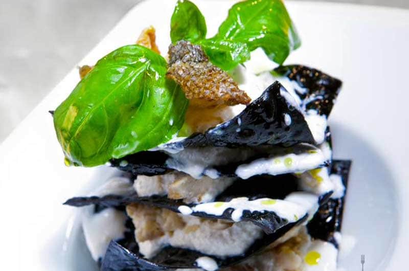 Triangoli di pasta nera disposti a strati con crema bianca, foglie di basilico e un elemento fritto sopra.