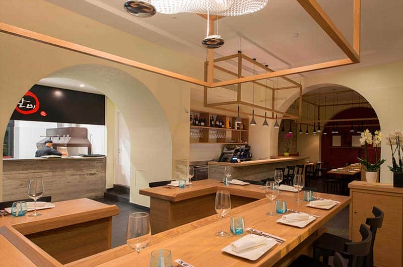 Tavoli in legno con bicchieri da vino e tovaglioli bianchi, sotto una struttura di travi in legno; a sinistra un arco apre su una cucina.