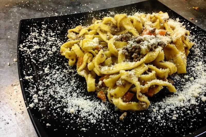 Tagliatelle con ragĂą di carne e carote, cosparse di formaggio grattugiato su piatto nero quadrato.