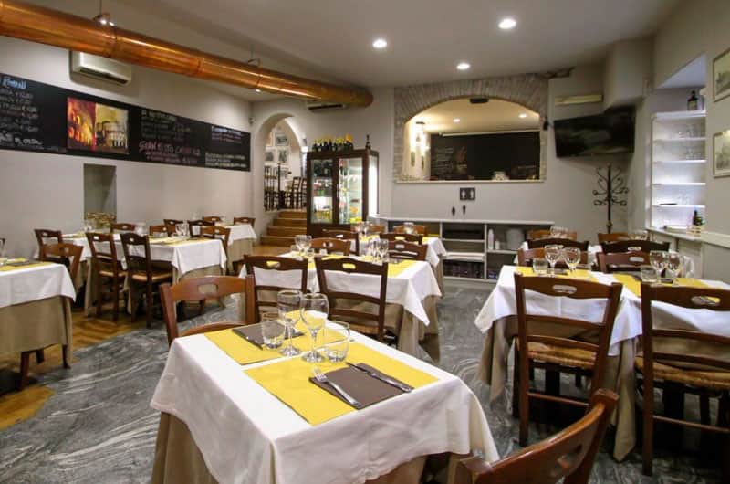 Sala da pranzo con tavoli e tovaglie bianche, tovagliette gialle e sedie in legno, arco in mattoni sullo sfondo.