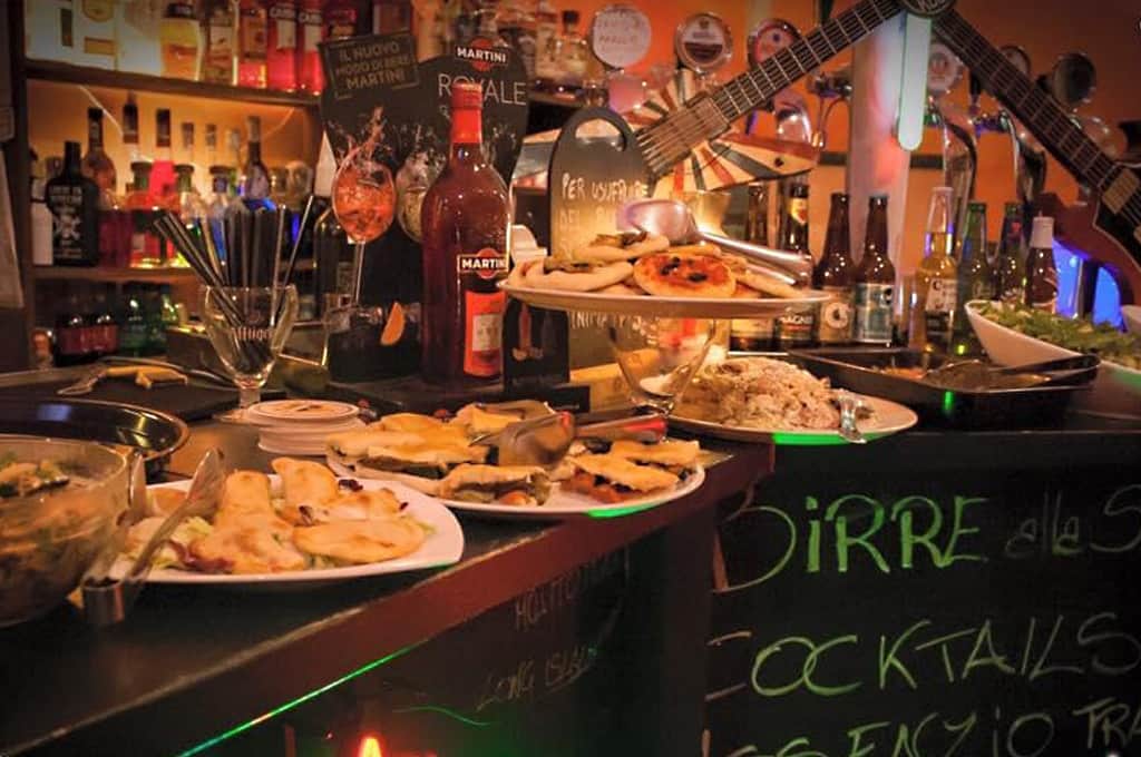 Bancone con vassoi di pizzette e riso, bottiglie di Martini, chitarre elettriche appese al muro e una lavagna con scritte in verde.