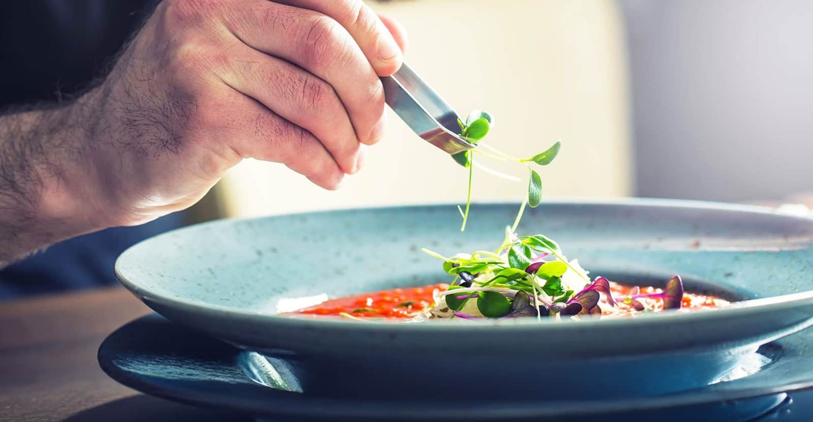Una mano guarnisce con pinzette da cucina un piatto di pasta corta con sugo rosso, erbe fresche e germogli.