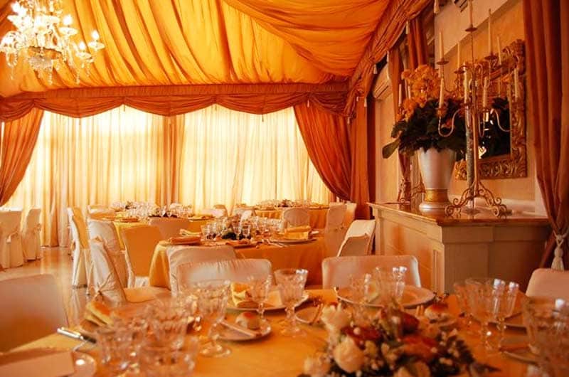Sala da banchetti con drappeggi arancioni al soffitto, tavoli rotondi apparecchiati e lampadario di cristallo.
