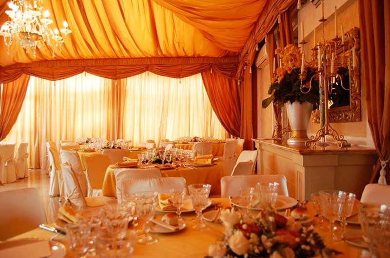 Sala da banchetti con drappeggi arancioni al soffitto, tavoli rotondi apparecchiati e lampadario di cristallo.