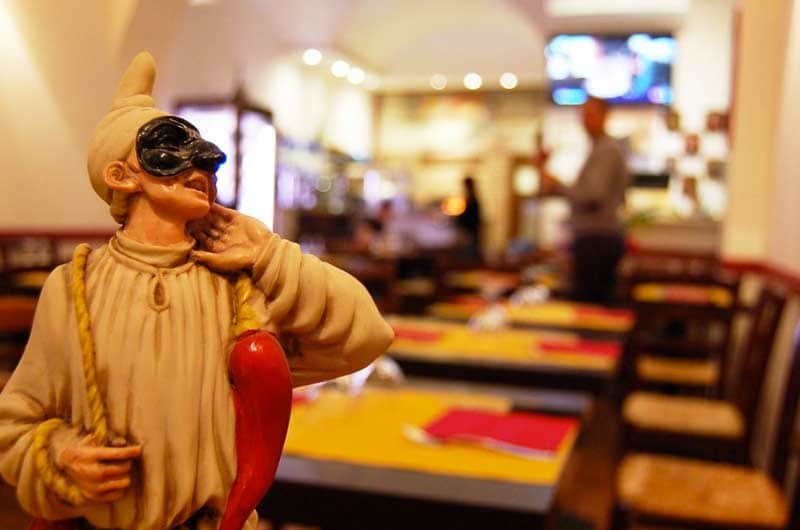 Statuina di Pulcinella con maschera nera in primo piano, sullo sfondo una sala con tavoli apparecchiati.