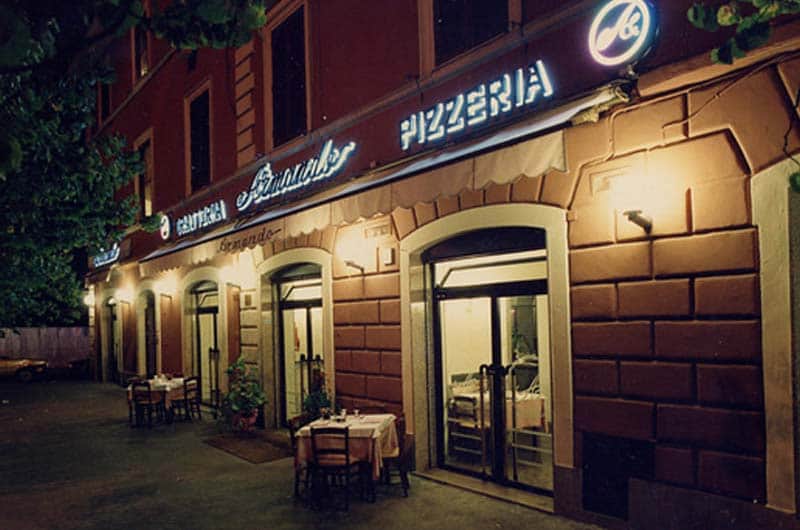 Facciata serale di un ristorante con archi illuminati, tavoli apparecchiati all'aperto e insegna luminosa con la scritta Pizzeria.