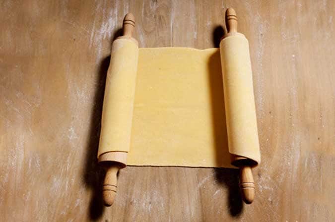 Sfoglia di pasta gialla arrotolata su due mattarelli di legno, su un piano infarinato.