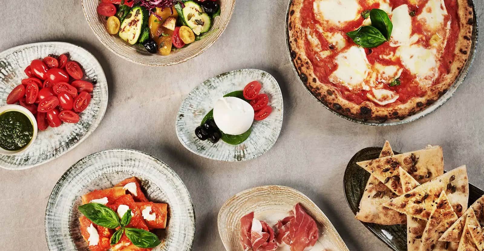 Vista dall'alto di una tavola con pizza margherita, burrata, pomodorini, salumi e triangoli di pane.
