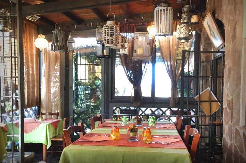Sala interna con tavoli apparecchiati di verde e rosso, lampade appese al soffitto e finestre sul dehors.