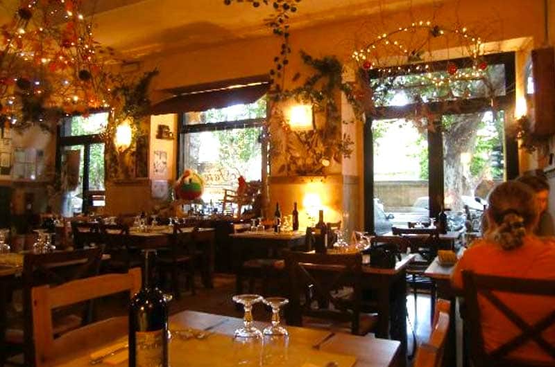 Sala di ristorante con tavoli in legno apparecchiati, luci decorative sospese dal soffitto e ampie vetrate su strada alberata.