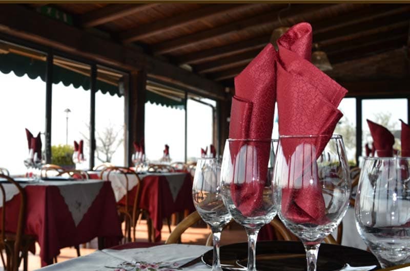 Bicchieri da vino con tovaglioli rossi piegati a punta, in una sala con travi di legno e vetrate.