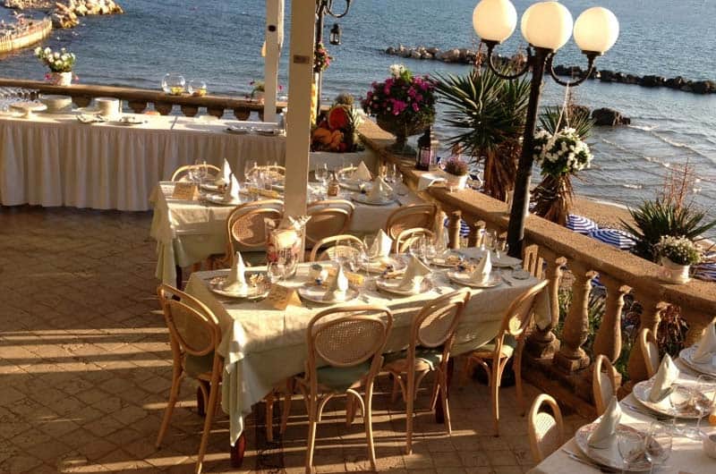 Tavoli apparecchiati su una terrazza affacciata sul mare, con lampioni e balaustra in pietra.