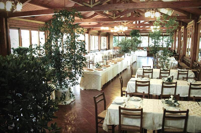 Sala da ristorante con travi in legno a vista, tavoli rotondi apparecchiati e piante alte tra gli spazi.