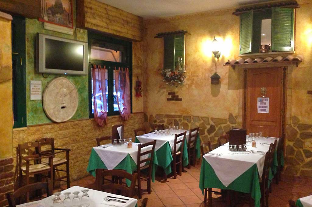 Sala con tavoli apparecchiati in verde e bianco, sedie in legno e pareti in pietra e intonaco giallo.