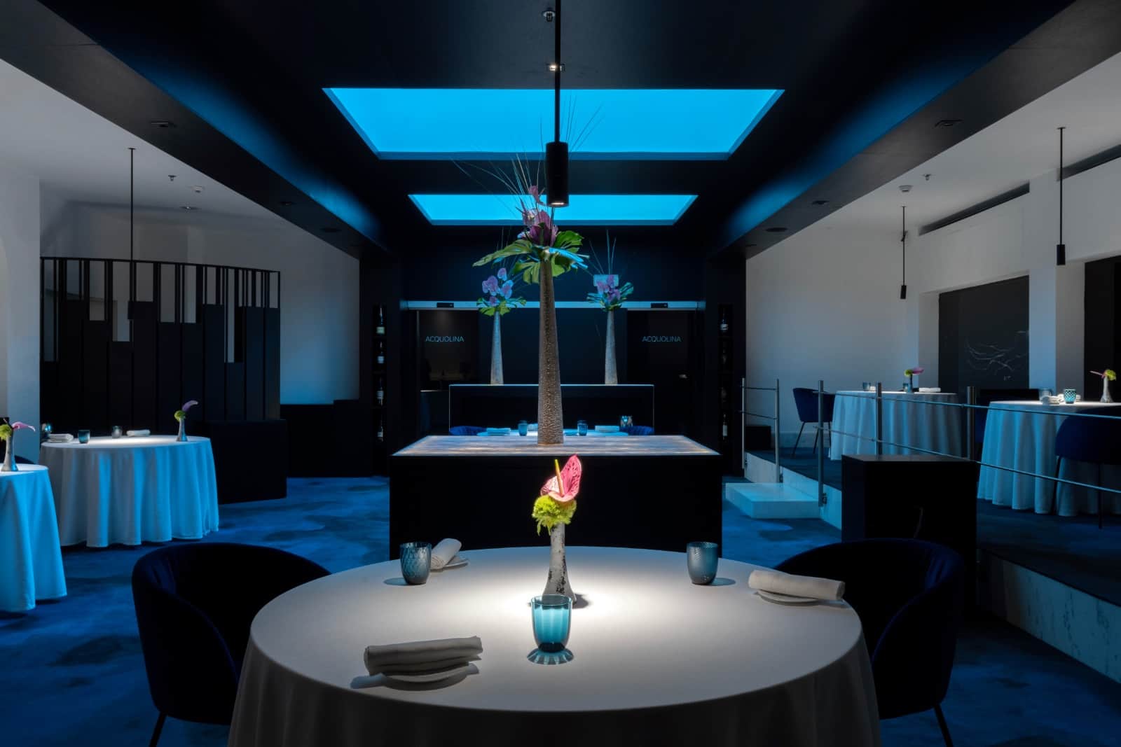 Sala blu con tavoli tondi apparecchiati, bancone centrale e insegna luminosa Acquolina sul fondo.