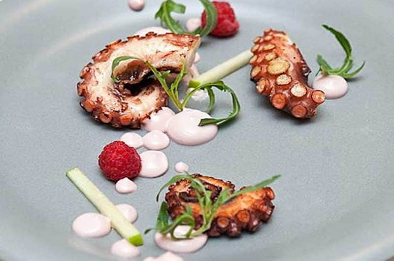 Tre pezzi di tentacolo di polpo grigliato su un piatto grigio, con lamponi, gocce di salsa rosa ed erbe verdi.