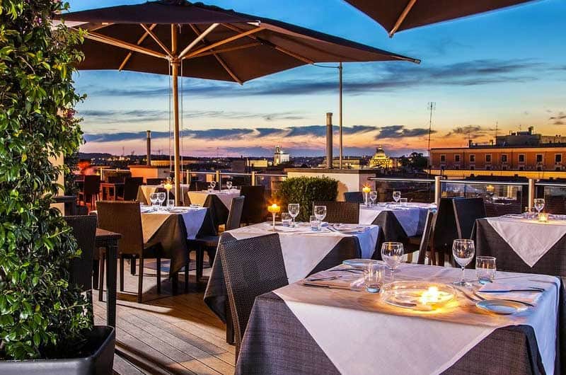 Terrazza con tavoli apparecchiati e ombrelloni, vista sui tetti di Roma al tramonto.