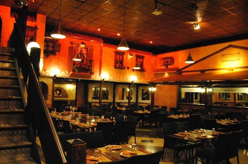 Sala da ristorante con luci calde arancioni, tavoli in legno e scala in metallo sulla sinistra.