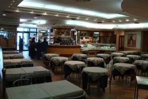 Sala di ristorante con tavoli quadrati apparecchiati in tovaglie chiare e bancone curvo sullo sfondo.
