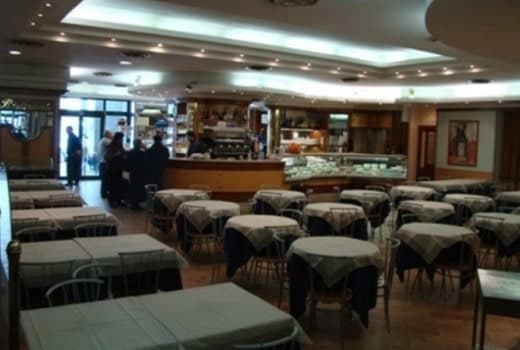Sala di ristorante con tavoli quadrati apparecchiati in tovaglie chiare e bancone curvo sullo sfondo.