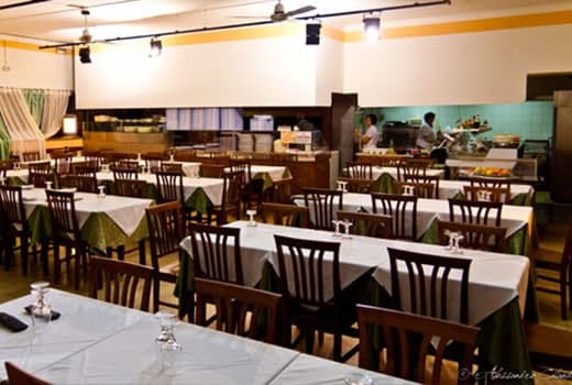 Sala da pranzo con file di tavoli apparecchiati con tovaglie bianche e verdi, sedie in legno scuro e cucina a vista sul fondo.
