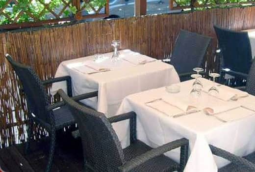 Tavoli con tovaglie bianche e sedie in vimini scuro su una veranda con recinzione di bambù.