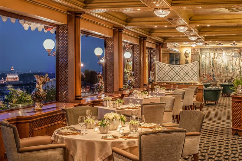 Sala da ristorante con tavoli rotondi apparecchiati, finestre ad arco affacciate su una veduta notturna della città con una cupola.