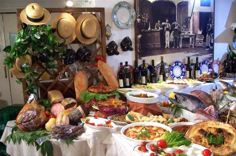Tavolo imbandito con salumi, pane, insalate, un pesce intero e bottiglie di vino, cappelli di paglia appesi sullo sfondo.