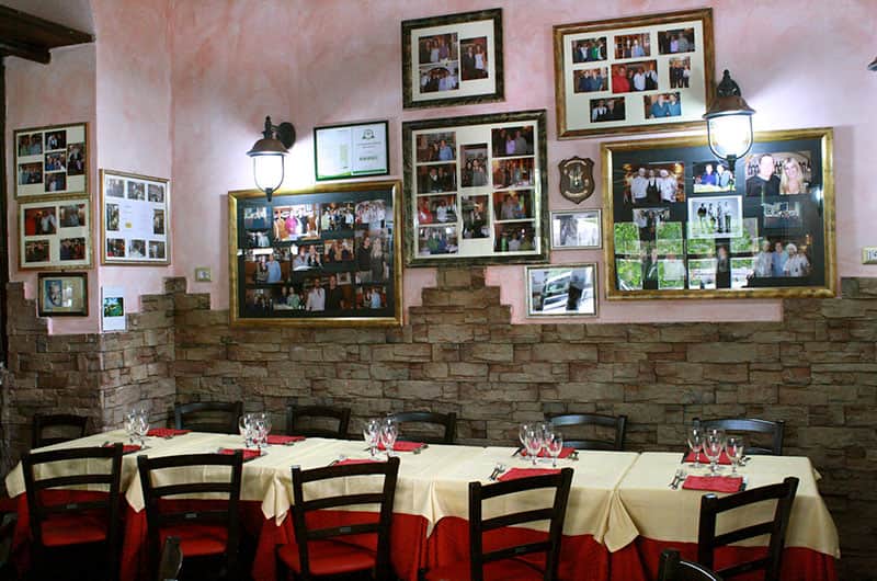Sala da pranzo con parete in pietra a vista e cornici fotografiche, tavolo lungo apparecchiato con tovaglia bianca e tovaglioli rossi.