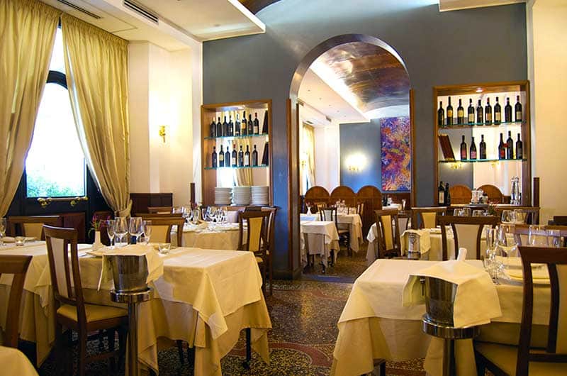 Sala da ristorante con tavoli bianchi, scaffali di vini ai lati di un arco centrale e tende dorate su una finestra.