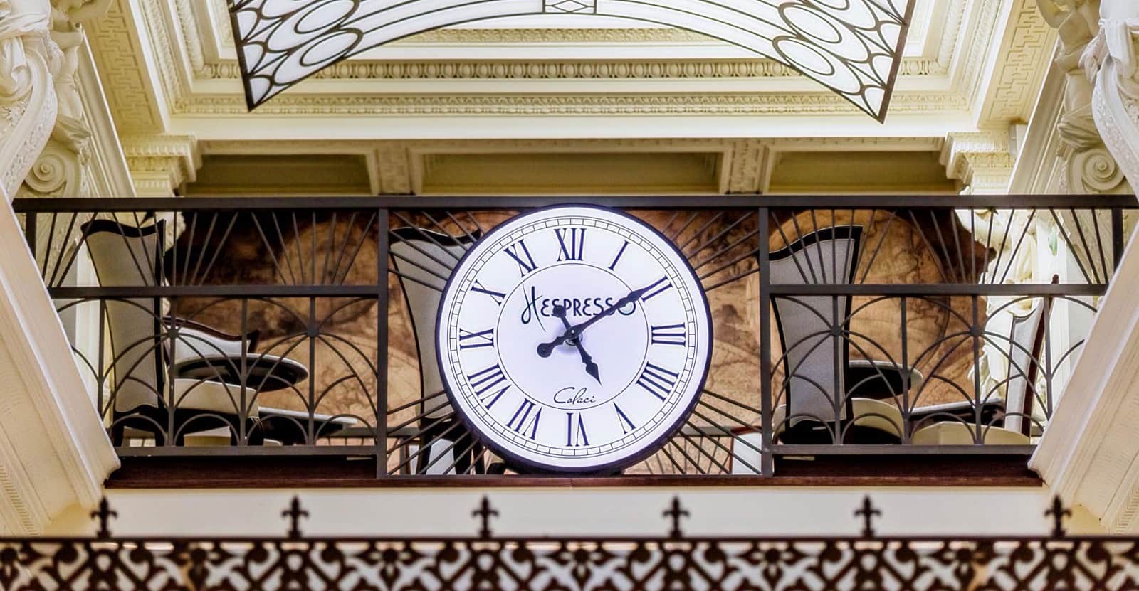 Grande orologio illuminato con numeri romani e scritta Hespresso, sospeso sotto un soffitto decorato.