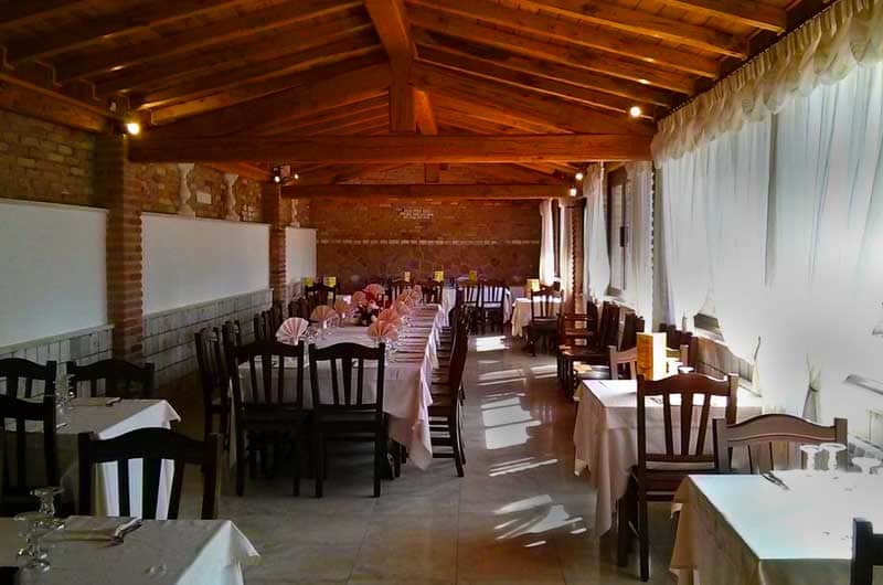 Sala da pranzo con soffitto in travi di legno, tavoli lunghi apparecchiati con tovaglie bianche e sedie in legno scuro.