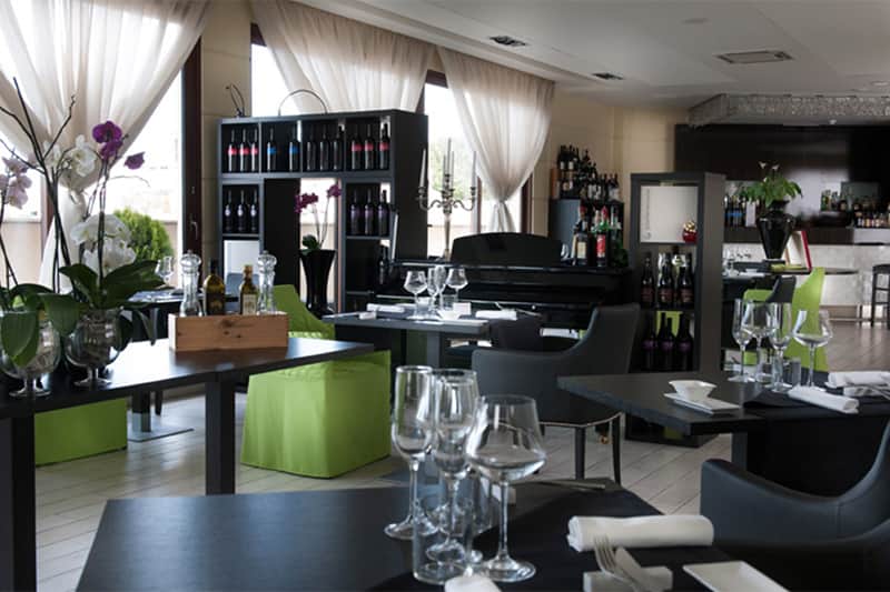 Sala di ristorante con tende bianche alle vetrate, tavoli apparecchiati e poltrone verdi.